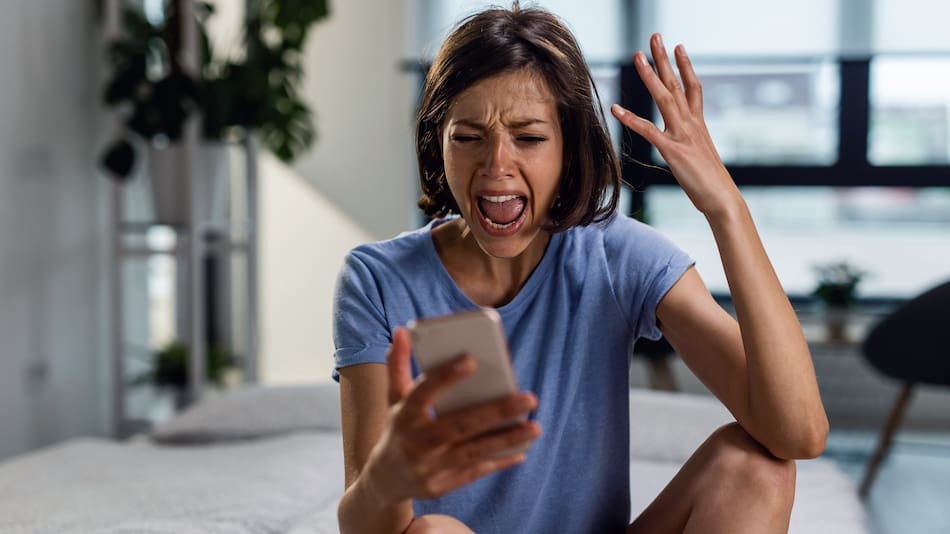 Frustrated woman shouting at her phone while sitting on the bed and reading text message she has received.